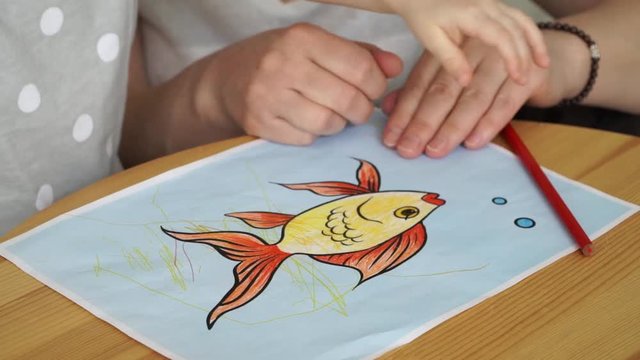 Close up of women's hands and children's hands draw goldfish with colored pencils on sheet of paper. Grandmothers and granddaughter