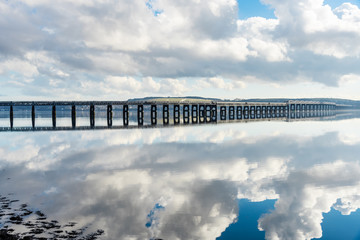 Railway bridge over a river on a partly cloudy winter day. a hillside town is visible in background. Reflection inwater. Firth of Tay, Dundee, Scotland.