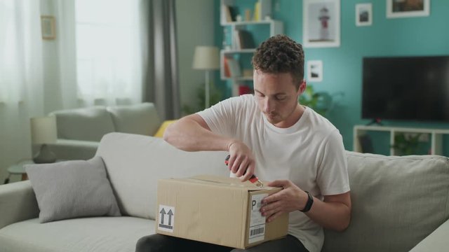 Young man sitting on couch in his living room and opening cardboard box package