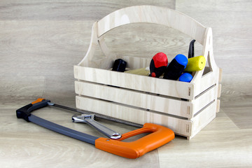 Wooden tool box with a handle, which contains a screwdriver, hammer and other tools. In the foreground is a hacksaw with an orange handle.
