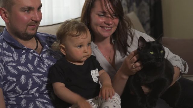 Family values: Mother and father playing with baby child boy indoor - Huge black panther like cat kitty - Parents wearing blue and gray home wear and their son black and white - Happy smiling people