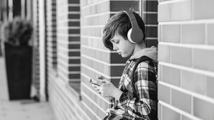 Teenage boy listening to music outdoors. Stylish boy using phone in urban setting. Happy teenage boy wearing wireless headphones, playing games on phone © volurol