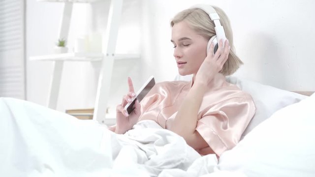 happy blonde girl touching headphones while listening music and looking at smartphone in bedroom