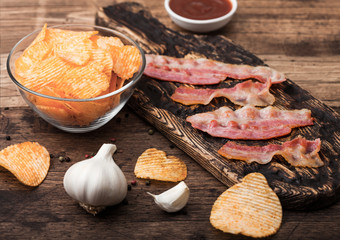 Potato crisps with backon flavour with grilled bacon rashers on vintage chopping board with garlic and sauce on wooden background.