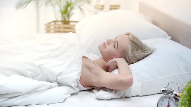 attractive blonde girl with closed eyes lying in bed, stretching and smiling in bedroom 