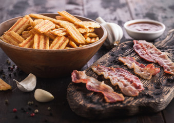 Bacon flavoured snacks chips with grilled bacon rashers on vintage chopping board with garlic and sauce on wooden background.