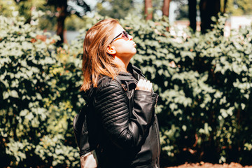 Fototapeta na wymiar Young woman with brown hair wearing sunglasses and a leather jacket sitting in the bright hot sun – Thirsty and tired girl on a torrid summer day – Concept image for dehydration and overheating