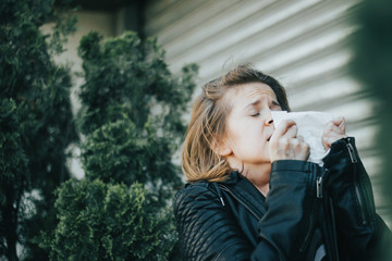Fototapeta premium Young woman with brown hair wearing a leather jacket holding a white napkin while sneezing – Girl having an allergic reaction or a virus infection – Concept image for allergy and sickness
