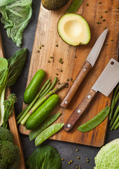Assorted green toned raw organic vegetables in wooden box on dark background. Avocado, cabbage, cauliflower and cucumber with trimmed beans and chopping board with knife.