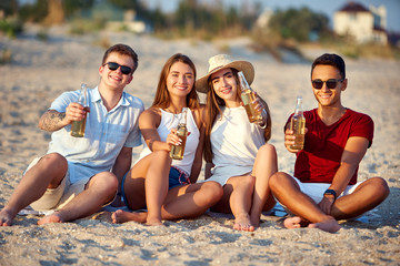Group of friends having fun enjoying a beverage and relaxing on the beach at sunset in slow motion. Young men and women drink beer sitting on a sand in the warm summer evening.
