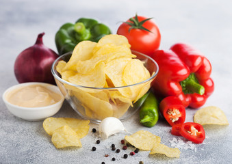 Glass bowl plate with potato crisps chips with onion flavour on light table background. Red and green chilli pepper with paprika and garlic with red onion and hot spicy sauce.