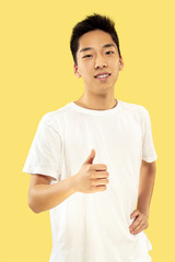 Korean young man's half-length portrait on yellow studio background. Male model in white shirt. Smiling and showing the sign of OK. Concept of human emotions, facial expression.