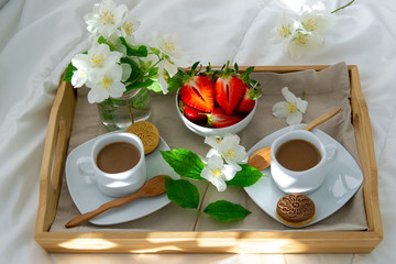 Breakfast in bed for two. Wooden tray with coffee, strawberries and cookies. Jewelry with delicate white flowers. Beautiful sunlight.