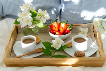 Breakfast in bed for two. Wooden tray with coffee, strawberries and cookies. Jewelry with delicate white flowers. Beautiful sunlight.