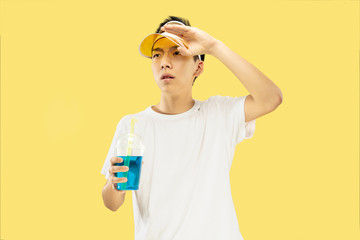 Korean young man's half-length portrait on yellow studio background. Male model in white shirt and yellow cap. Drinking cocktail. Concept of human emotions, expression, summertime, vacation, weekend.
