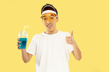 Fototapeta na wymiar Korean young man's half-length portrait on yellow studio background. Male model in white shirt and yellow cap. Drinking cocktail. Concept of human emotions, expression, summertime, vacation, weekend.