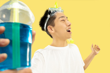 Korean young man's half-length portrait on yellow studio background. Male model in white shirt and swimglasses. Drinking cocktail. Concept of human emotions, expression, summertime, vacation, weekend.
