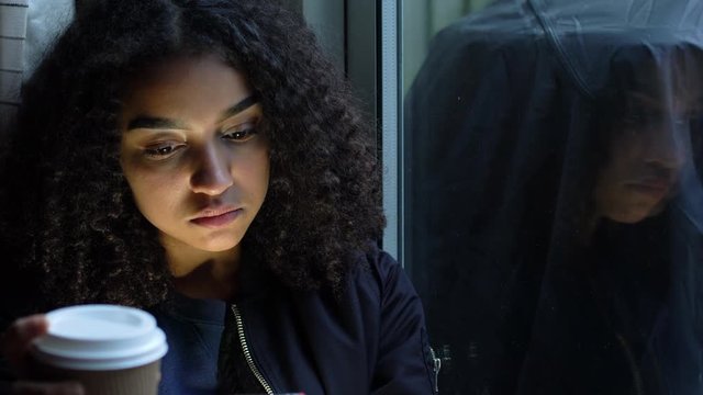 Sad depressed beautiful female mixed race African American girl teenager young woman teen wearing a black jacket sitting using her mobile cell phone looking out of a window and drinking cup of coffee