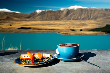 Cup of hot chocolate and smoke salmon sanwich on cement table with mountain deep blue lake view at...