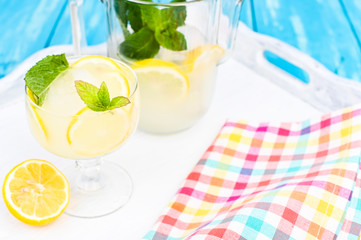 Lemonade with ice, lemon slices and mint in a glass and a carafe on a blue wooden table. Summer cold refreshing drink with fruits and vitamins.