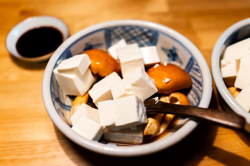 Fototapeta na wymiar Traditional japanese ceramic bowl in restaurant with wooden table and spring vegetable dish with honey mushrooms and cubed tofu with soy sauce