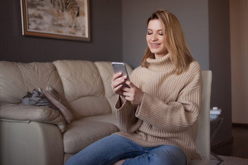 young attractive woman in a sweater sits at home in the living room and uses a smartphone.