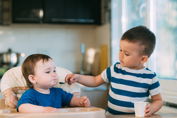 Kids in the kitchen, brother feeds baby sister yogurt with a spoon, she helps parents