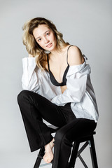Fashion model sitting on a chair in a white blouse and flared pants barefoot on a white background