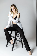 Fashion model sitting on a chair in a white blouse and flared pants barefoot on a white background
