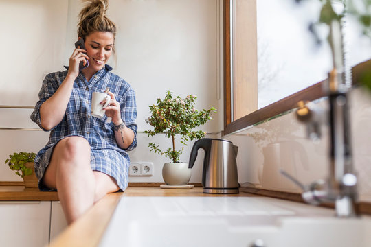 beautiful and young woman having breakfast at home and having fun, sitting on the kitchen and talking on the phone
