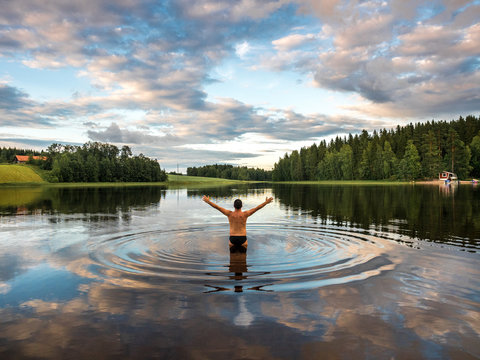 Back view of man standing in lake and opening arms while enjoying landscape with forest and houses in Finland