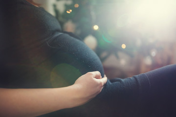 Detail of pregnant woman sitting in front of Christmas tree holding hands on her belly.