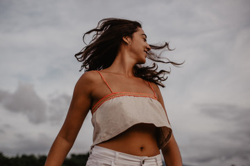 Side view of excited young female smiling and dancing with closed eyes and raised hands near pond with calm water in countryside