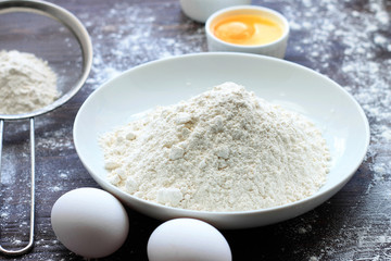 Flour powder sprinkled on wood table, eggs, sieve, table filled with flour.