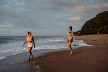 Two slim young females in shorts and bras smiling and looking at each other while standing on sandy shore against cloudy gray sky