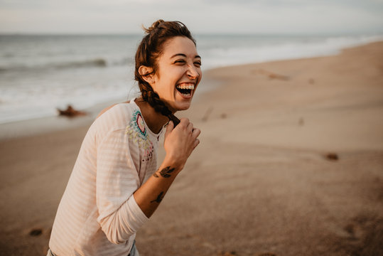 Young lady in casual outfit touching braid and laughing out loud while standing on sandy seashore