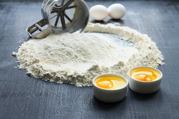Mug sieve flour and eggs. Mug sieve is on the flour, flour is scattered around on dark background.