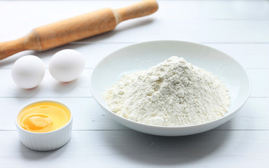 Fototapeta na wymiar A plate with flour, eggs, a rolling pin on a white wooden background. Pancakes ingredients. Egg in the bowl.