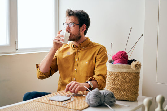 Adult male enjoying fresh hot tea and looking away while sitting at table near knitting needles and yarn