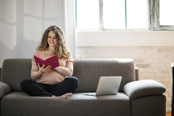 Beautiful pregnant woman working behind a laptop sitting on the sofa. The happiest time for every woman.