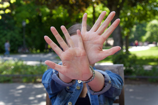 Junge Frau in einem Park sitzend h&auml;lt beide H&auml;nde abwehrend vor ihr Gesicht,