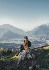 girl in an orange jacket and hat sits on a stone with her back and looks at the mountains