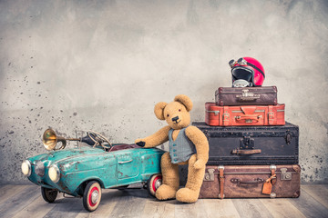 Teddy Bear toy standing near rusty retro  pedal car from 60s, antique travel trunks luggage, old leather valises, red helmet with outdated goggles front loft background. Vintage style filtered photo