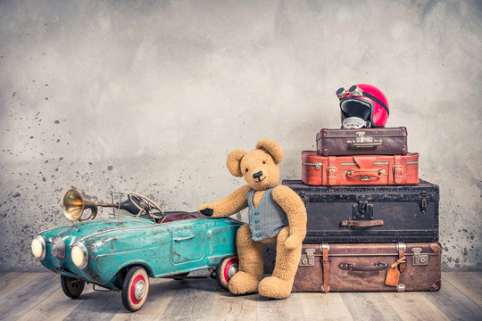 Teddy Bear toy standing near rusty retro  pedal car from 60s, antique travel trunks luggage, old leather valises, red helmet with outdated goggles front loft background. Vintage style filtered photo
