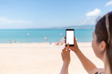 Woman holding up a smart phone at beach. Sea, sand and sky on the background. Concept of modern technology and internet. Copy space and mock up