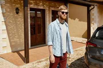 Horizontal shot of trendy looking young bearded male traveler in his twenties standing by car at open trunk, carrying suitcase, posing outdoors with nice guest house in background, looking at camera