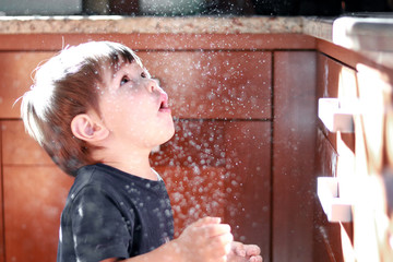 Little playful boy with surprised face expression playing with flour throwing it up at kitchen at sunlight. Happy childhood.