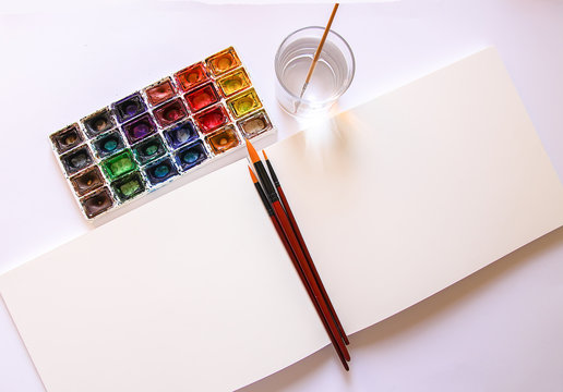 Watercolor paints, brushes, water in glass and paper on white background. Flat lay, top view