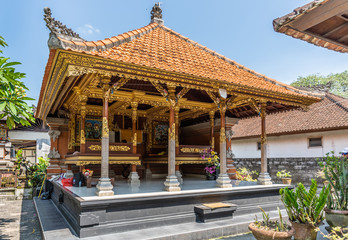 Dusun Ambengan, Bali, Indonesia - February 25, 2019: Family compound. Open wall Hundu temple with large beds in front of altar. Browns and lots of gold, blue sky, green vegetation, red roof.