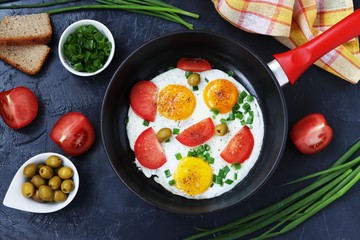 Scrambled eggs with tomatoes and green onions in a frying pan on a dark background, top view, on the table are olives, bread and tomatoes